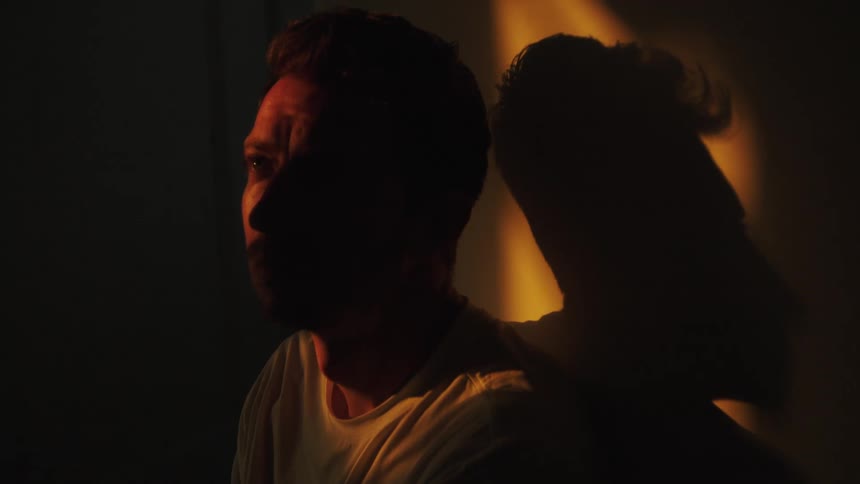 The video shows a closeup of a person sitting in the dark dramatically lit by a warm orange light from the right which creates a large dark shadow of their profile on the wall behind them the figure is dimly visible in profile appearing to