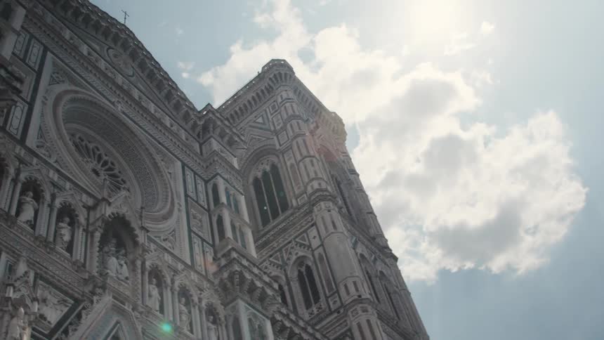 The video is an extreme lowangle upwardpanning shot of the white and green marble facade of the florence duomo complex likely the cathedral or baptistery the architecture is strikingly detailed and is dramatically framed against a bright