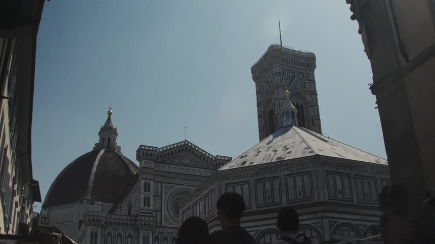 The video captures an upward view of the florence duomo complex santa maria del fiore battistero and giottos campanile from a narrow street the iconic dome and striped bell tower dominate the frame against a bright clear sky