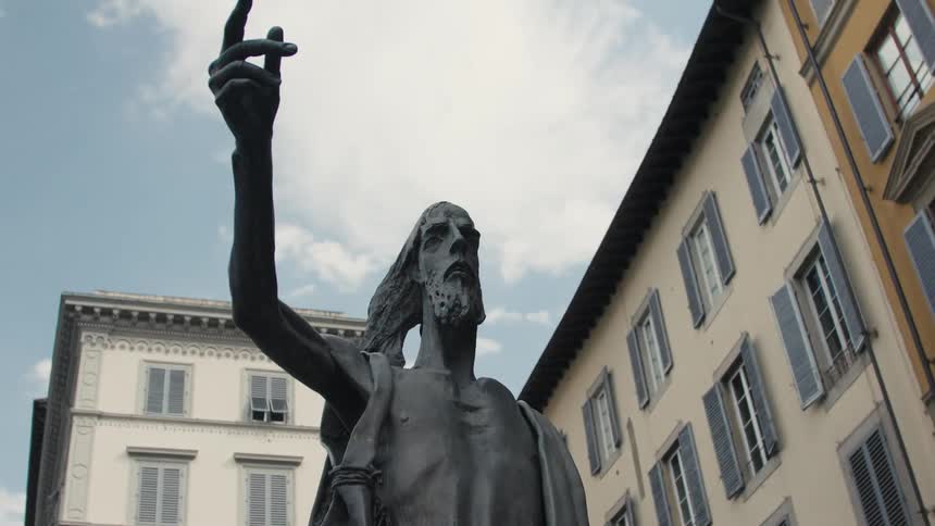 Statue of a bearded figure with raised arm in a square surrounded by old buildings