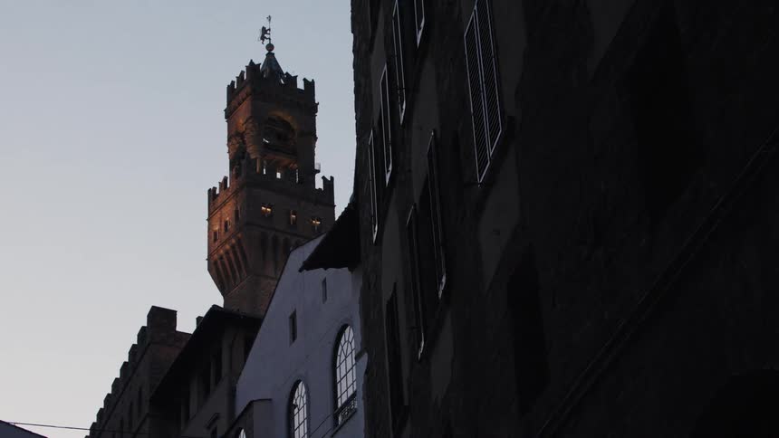 The video presents a striking view of a tall medievalstyle bell tower with battlements illuminated from within at dusk or twilight the tower is framed between the dark imposing silhouettes of narrow european city buildings in the foreground