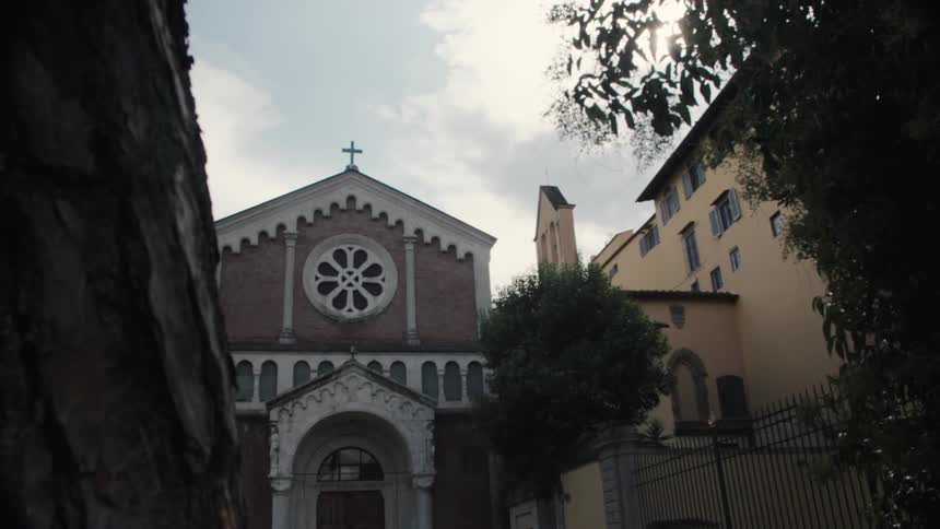 The video is an exterior upwardangled shot of an old church facade with a prominent rose window and a cross at the very top framed by dark foliage and an unidentifiable stone structure in the immediate foregroundthe sun shines brightly from