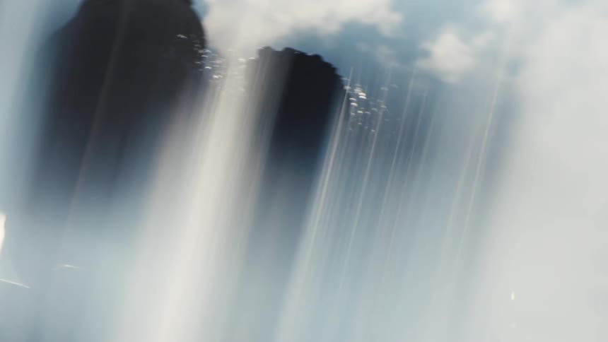 The video is an extreme closeup shot from the bottom of a massive waterfall where thick white sheets of water cascade down in front of dark craggy rock faces the bright sunlight creates an atmospheric effect with the heavy mist and spray f