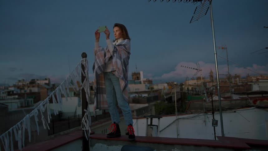 A woman on a rooftop at dusk calmly holding up her phone to film wrapped in a poncho and jeans with reddotted boots as pennants flutter over the city behind her