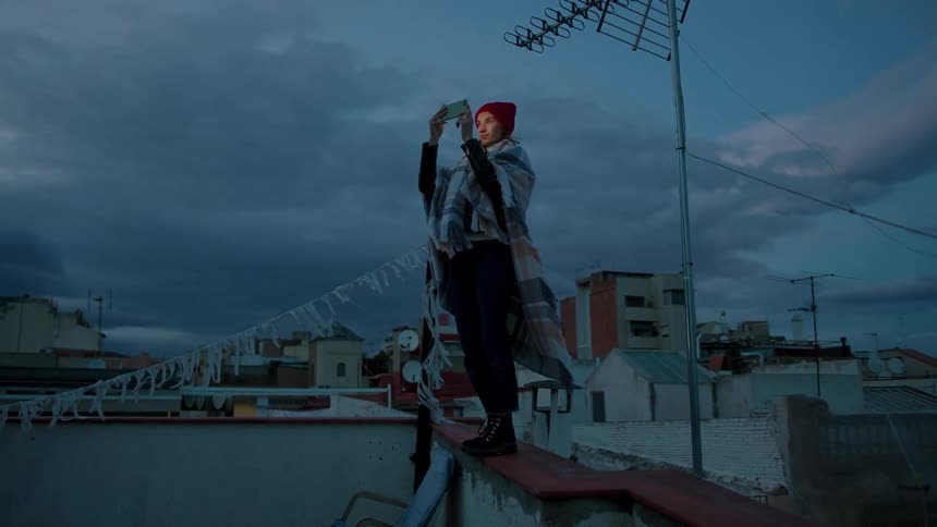 Woman in poncho and red beanie holds phone above head on building ledge at sunset