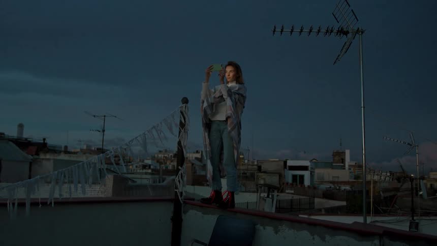 Woman standing on a ledge at dusk holding a phone and taking a photo on top of a building