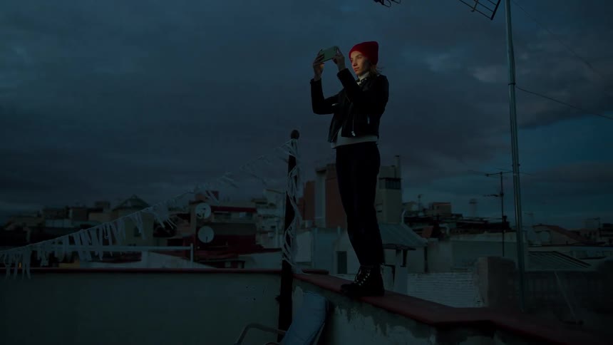 Young woman wearing a red beanie standing on the ledge of a building filming on her smart phone outdoors during dusk or dawn