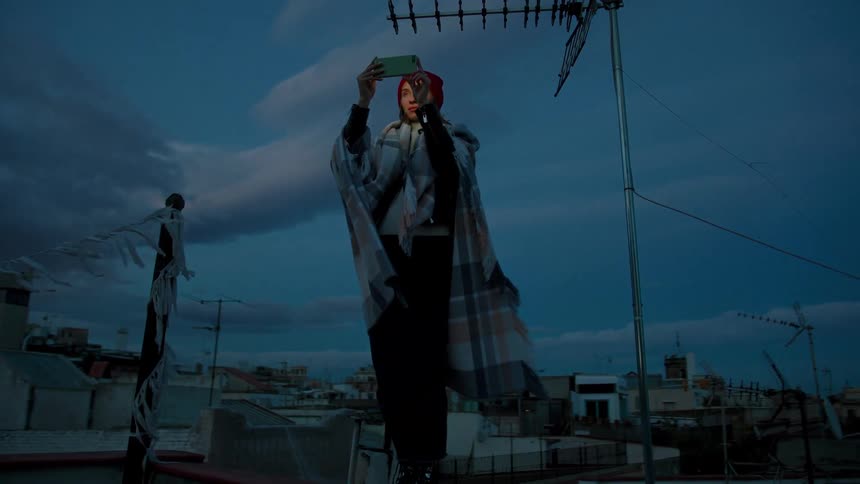 Woman wearing a beanie and blanket takes a photo with her phone on a rooftop at dusk