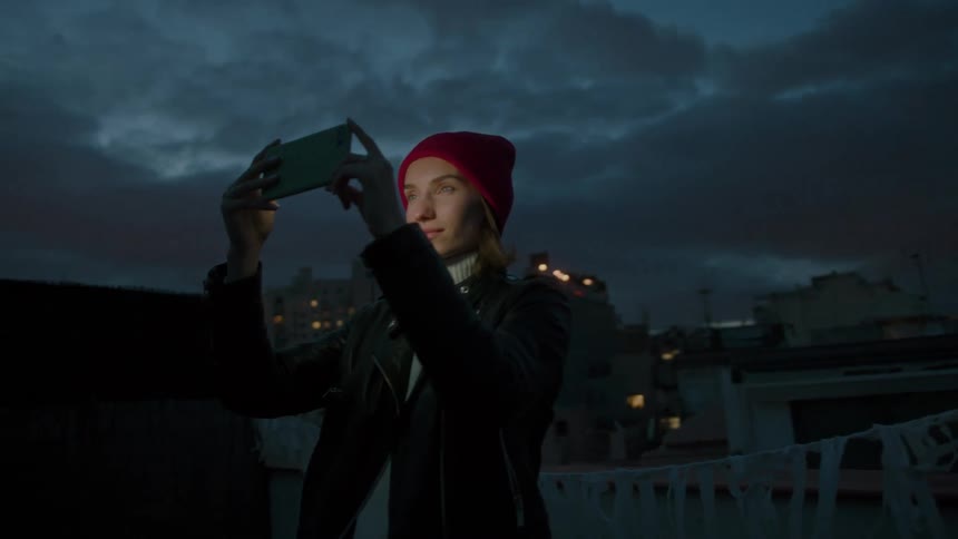 A young woman wearing a red beanie and a black jacket stands on a rooftop terrace at dusk or night holding a green smartphone up to record or take a picture of the city buildings and cloudy sky around her