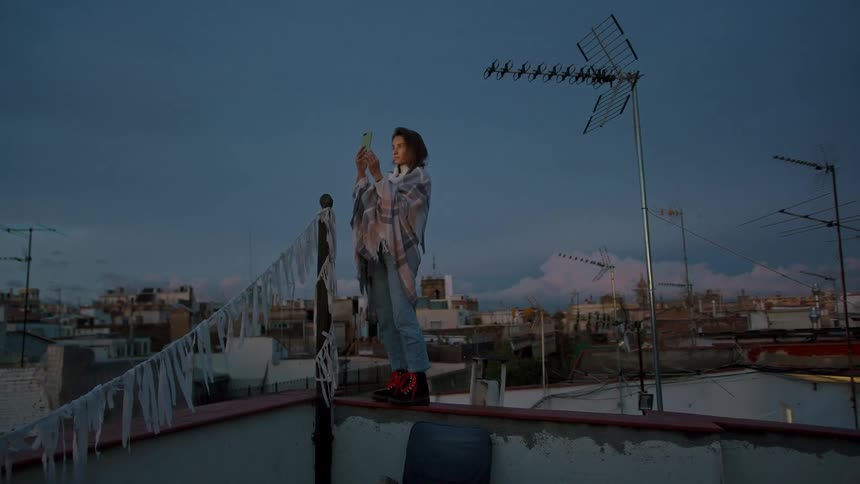 A woman stands on a rooftop parapet at dusk serene and focused as she films with her phone wrapped in a plaid shawl and jeans amid antennas and pinktinged clouds over the city