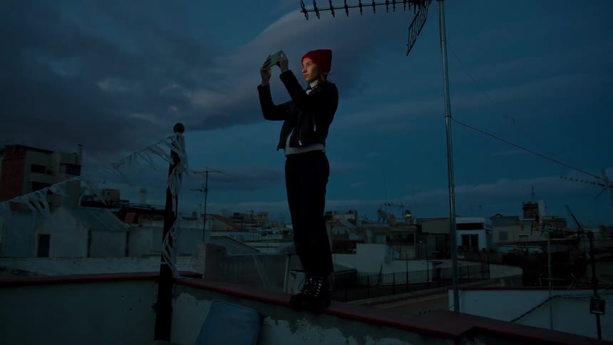 Woman wearing a red beanie standing on a ledge at dusk taking a photo of the city with her phone