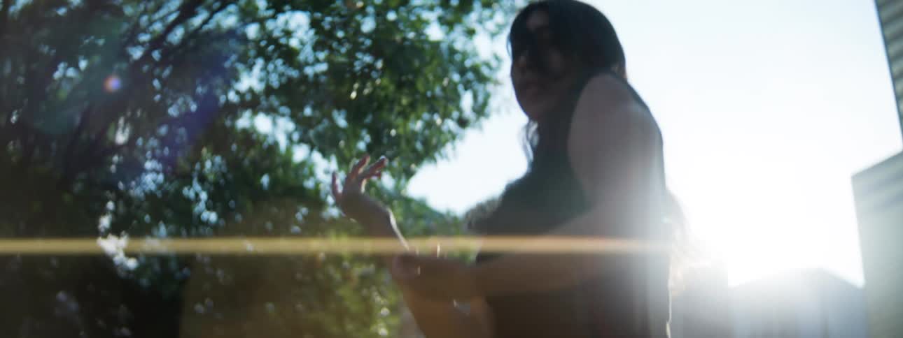 Woman raises her arms amid sunlight filtered through trees and buildings in the background