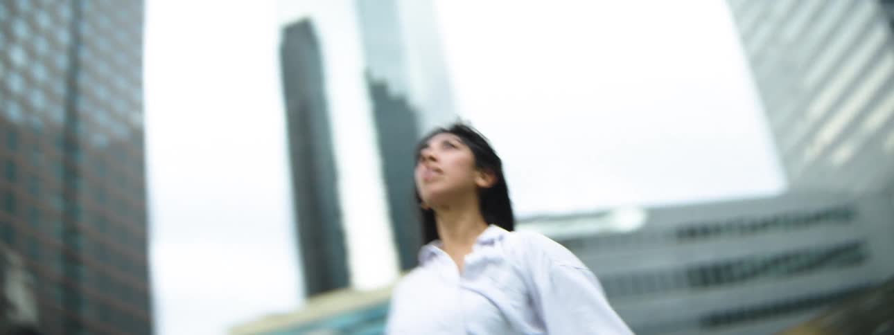 A woman with long dark hair performs dynamic expressive modern dance movements in a park setting raising her arms and spinning with intensitythe scene contrasts her flowing white shirt and uninhibited movement with the modern towering glass building