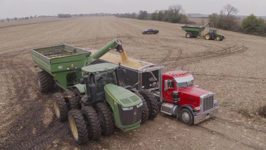 The video is a wide aerial shot of a tractor and large grain cart pulled up next to a red semitruck in a harvested field the tractor is actively transferring grain from the carts auger into the trucks trailer