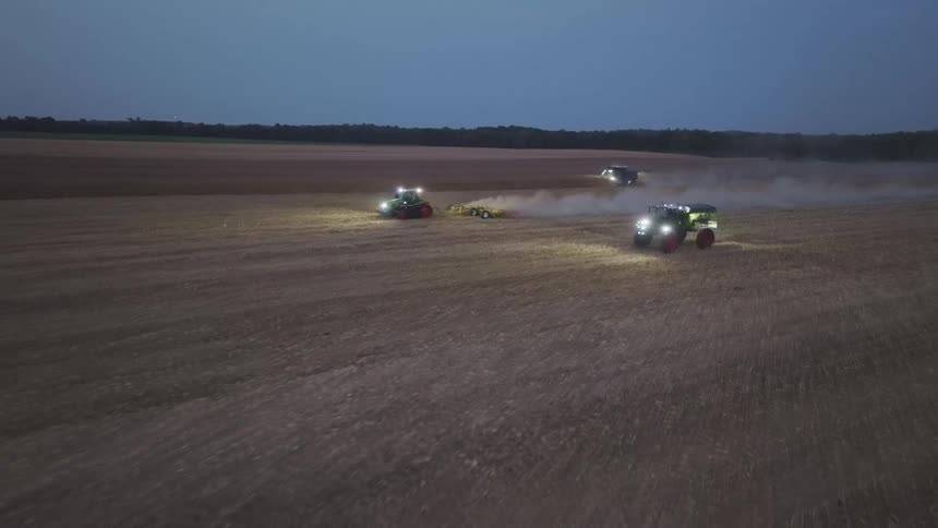Tractors working in an agricultural field at dusk with lights on and dust in the air