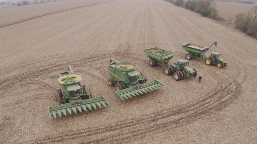 An aerial view shows a wide stubble field with a neat lineup of green farm machines including two combines with corn heads two grain carts and a tractor tire tracks looping around and a few people standing nearby