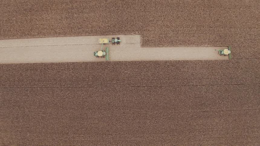 Overhead multiple green farm machines including combines tractors and grain carts work in a straight cleared lane that cuts through a vast brown field leaving precise parallel rows behind