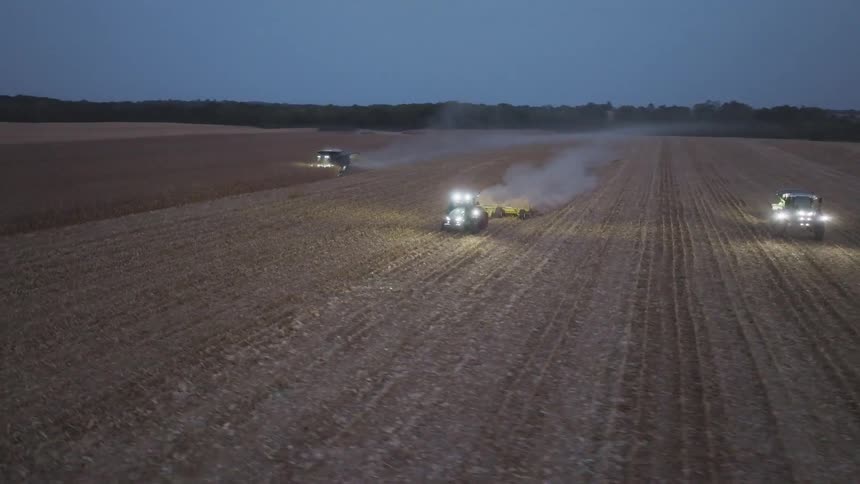 An aerial view video shows three large farm tractors with their headlights illuminated working in a wide harvested field after dark stirring up dust and smoke into the dim twilight air