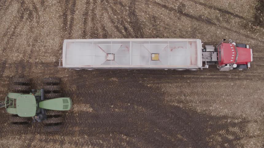 The video is a topdown aerial shot of a semitruck trailer parked in a harvested field of dirt and crop stubble a green auger wagon pulled by a tractor moves into the frame and prepares to unload grain into the trucks hopper