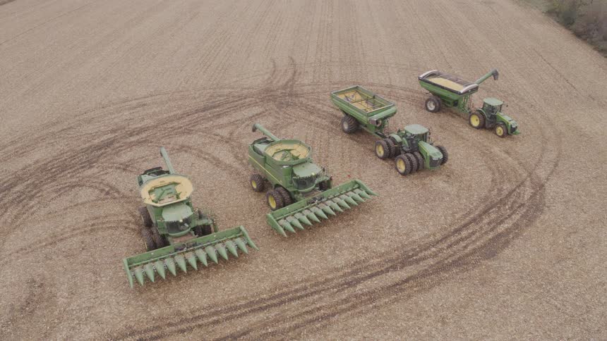 The video is a stationary aerial shot of a line of four modern farm vehicles two combines a tractor and an auger wagon parked diagonally on a recently harvested field of stubble the equipment primarily colored green and yellow takes up the