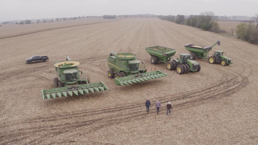 In a wide harvested field three people walk toward a lineup of green farm machines including two combines with corn heads two grain carts a tractor and a pickup with looping tire tracks curving across the stubble
