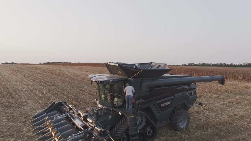 A man climbs into a tractor parked in a sunlit farm field at midday surrounded by open land and clear skies