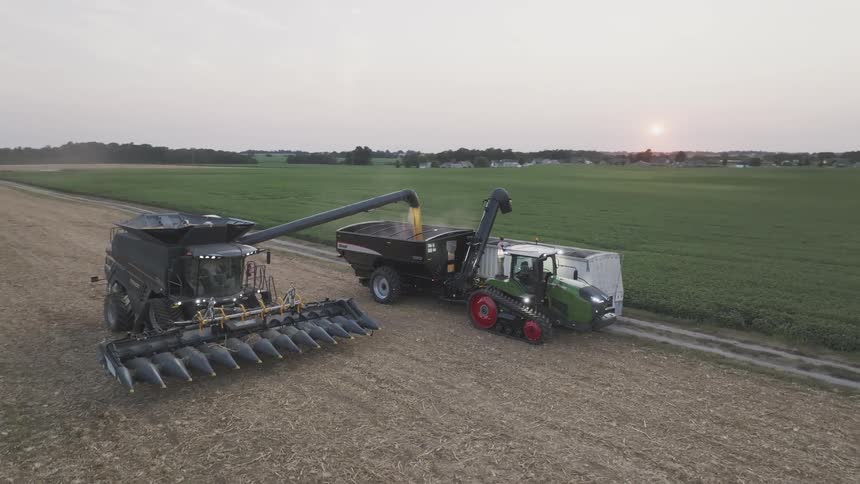 At sunset a combine harvester transfers its load of grain into a waiting grain cart pulled by a tractor while a second tractor loads a semitruck on a nearby dirt road