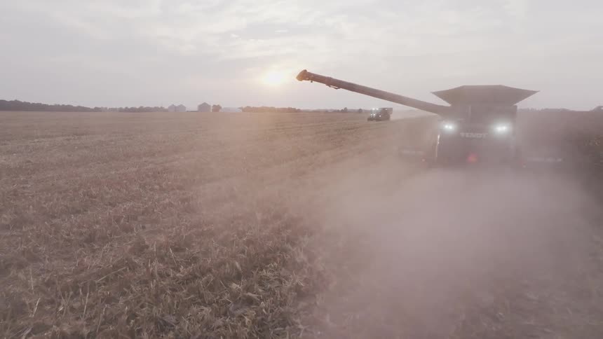 A combine harvester unloads corn into a tractor trailer across a dusty field