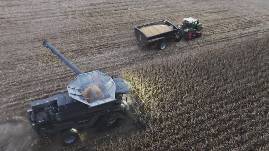 A high angle aerial view a combine harvester with its lights on moves through a field offloading its harvested grain into a tractor pulled cart driving alongside