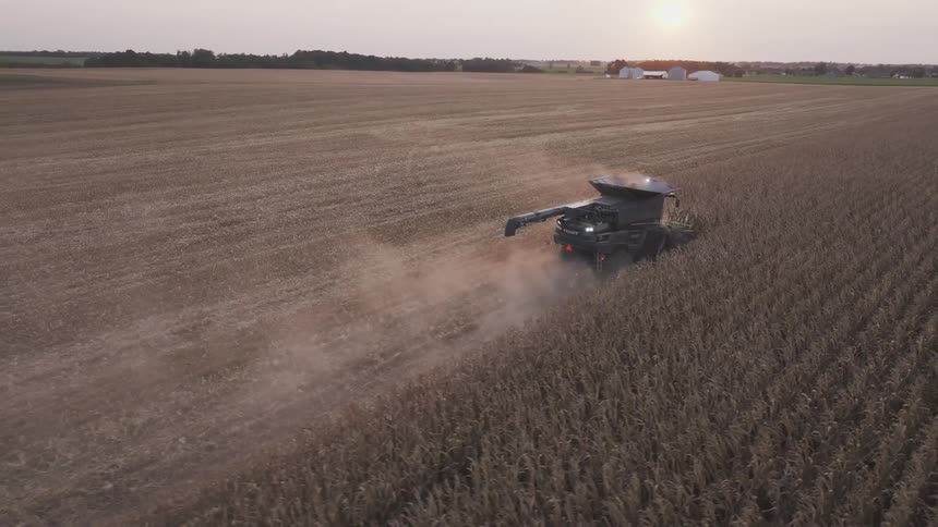 An aerial view shows a combine harvester cutting a dark swath through a vast brown cornfield trailing a plume of dust toward distant farm buildings
