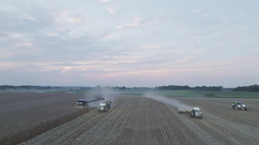 The video is a wide aerial shot of a field harvest in the early morning or late evening showing multiple tractors and combine harvesters working their bright headlights illuminate the rows of crops and the dust trails left behind against the dust