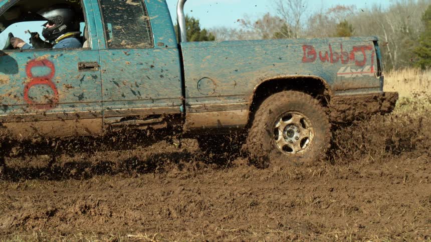 A mud splattered blue truck marked with the number 8 spins its front tire and flings clods as a helmeted driver grips the wheel during an off road churn