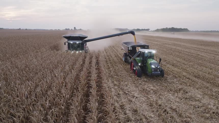 The video is a highangle shot of a modern combine harvester reaping a field of dry brown crops likely corn or grain at dusk a green tractor towing a large cart drives alongside the combine as it unloads the harvested grain into the cart v