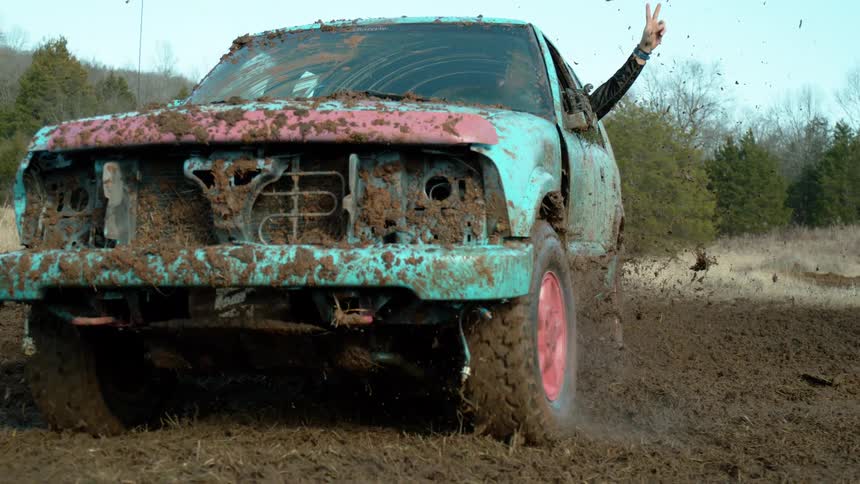 A mud spattered turquoise and pink sedan barrels through a churned field front bumper missing wheels flinging dirt as a passengers arm juts out the window flashing a peace sign