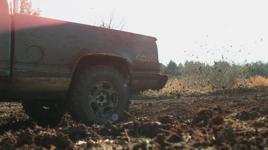The video shows an old mudsplattered pickup truck with a brush guard driving forward slowly in a deeply rutted dirt or mud field the low angle captures the intense backlight of the sun through the trees emphasizing the messy offroad envi