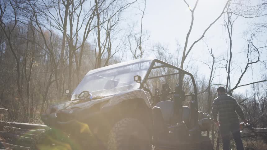 The video shows a man in a plaid flannel shirt and jeans standing beside an offroad utility vehicle utv holding a chainsaw at his side the setting is a bright sunlit forested area with a clearcut foreground littered with logs and tr