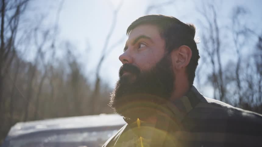 The video is an extreme closeup shot of a rugged man with a large beard looking up towards the sky in a wooded area the shot uses a lot of natural light and lens flare giving the scene a warm bright and cinematic feel