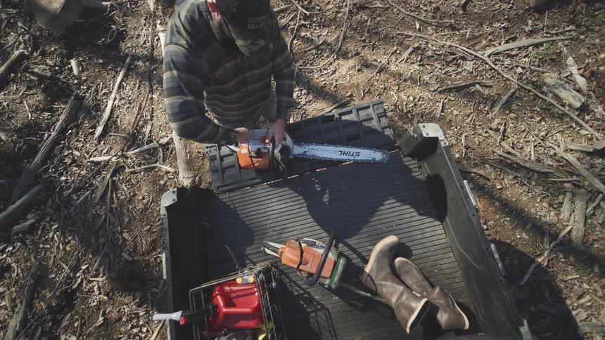 A man in a camo cap grabs a chainsaw from a truck work boots arranged in the bed amid a debris strewn logging site