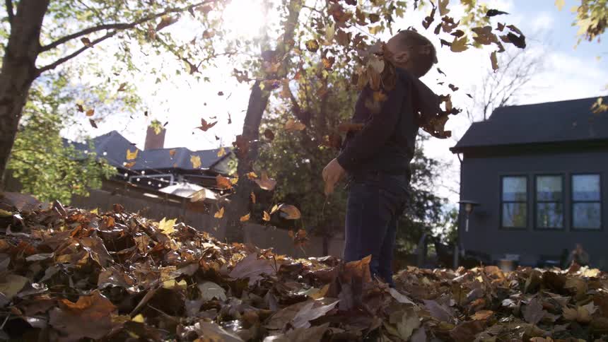 A kid wearing a hoodie stands in his backyard at midday scooping up a bunch of fallen leaves from the ground and tossing them into the air watching with excitement as they scatter around him