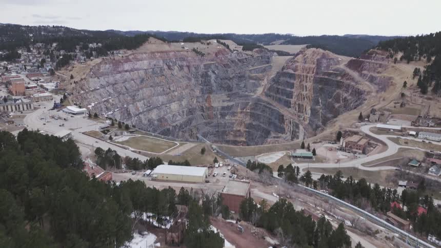 An aerial drone camera moves forward and upward over a forested hillside to reveal a vast terraced openpit mine excavation bordering a scattered settlement of buildings and roads beneath a cloudy sky