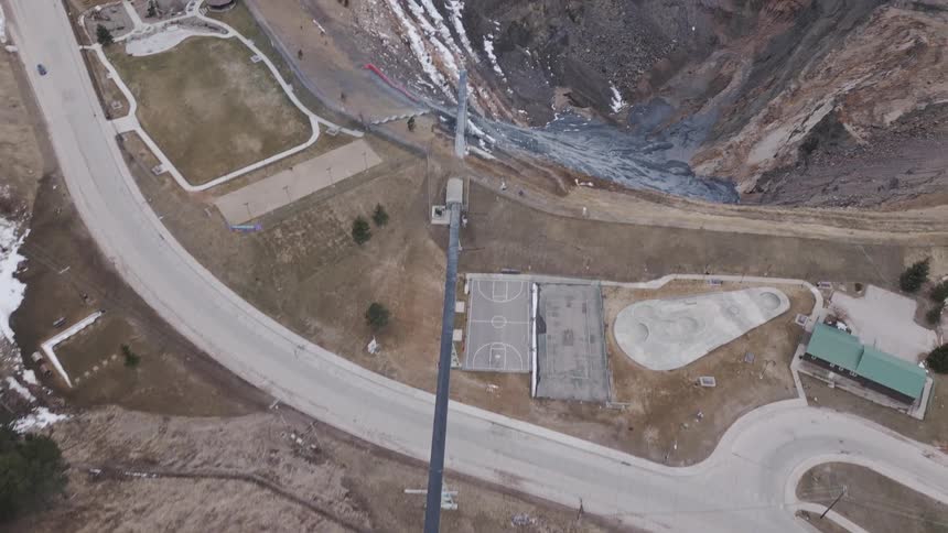 An aerial view follows a long conveyor up a hill before tilting to reveal a vast terraced open pit mine next to a small town