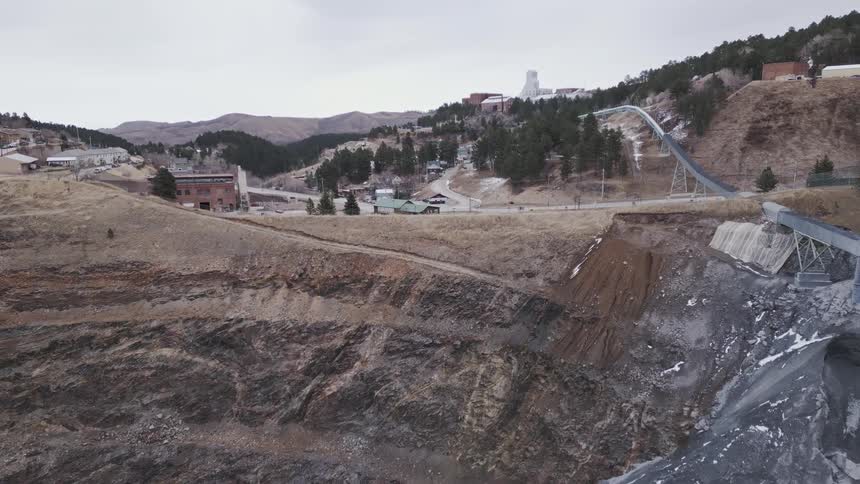 The video begins with a view of a large deep excavation possibly an old mine pit with a cascade of dark fine material flowing down one sideas the camera pans up and out it reveals a small town nestled in a valley featuring houses roads and a pr