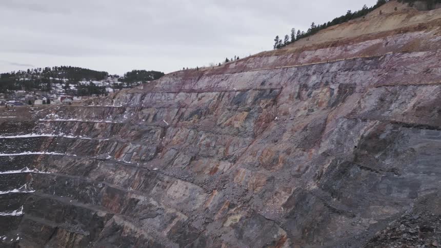 An aerial video slowly reveals the immense tiered and steep walls of a deep openpit mine showing multicolored geological strata and patches of snow with the rim topped by pine trees and a distant mining town in the background under a cloudy sky