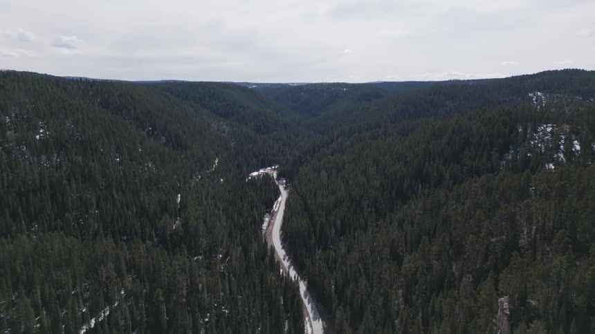 An aerial view shows a winding road cutting through a deep forested valleythe valley slopes are covered in dark evergreen trees with patches of snow all under an overcast sky