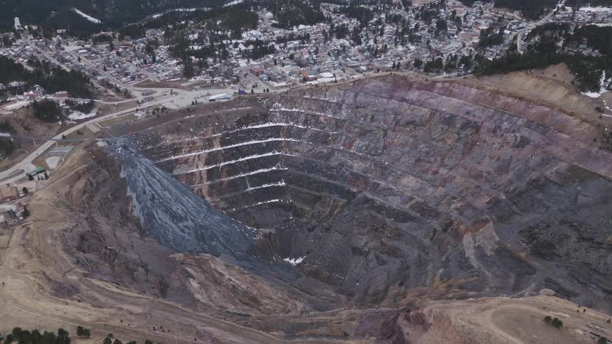 A high aerial shot reveals a huge terraced openpit mine with spiral haul roads bordered by a small town and forested snowspeckled hills