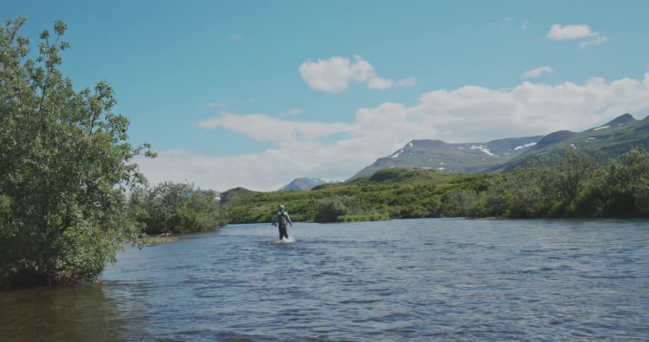 Fisherman walking through a river surrounded by vegetation with mountains in the background