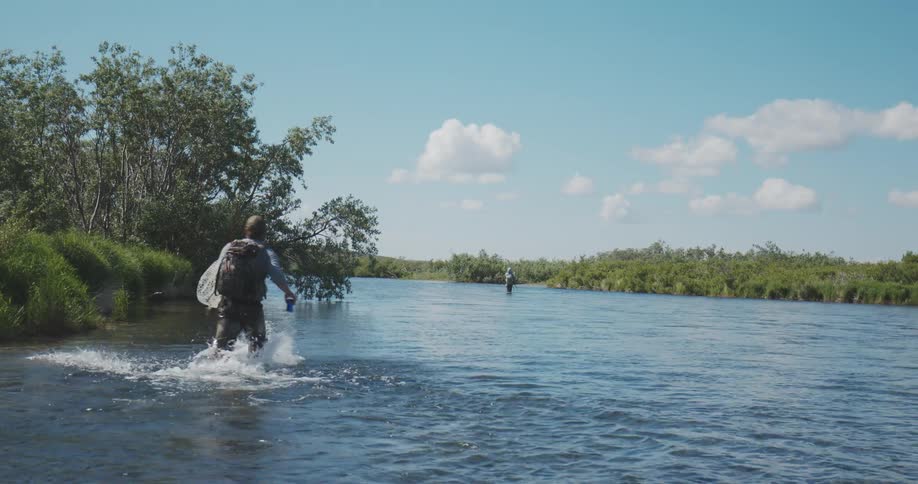 Two fly fishermen wade through a wide river on a sunny day surrounded by lush green riverbanks