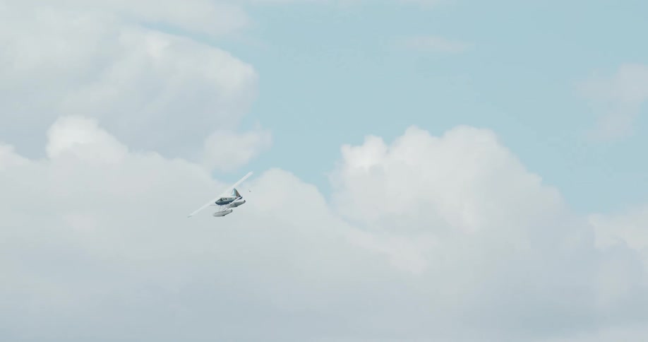 A small singleengine propeller aircraft banks left against a pale blue sky filled with towering white clouds