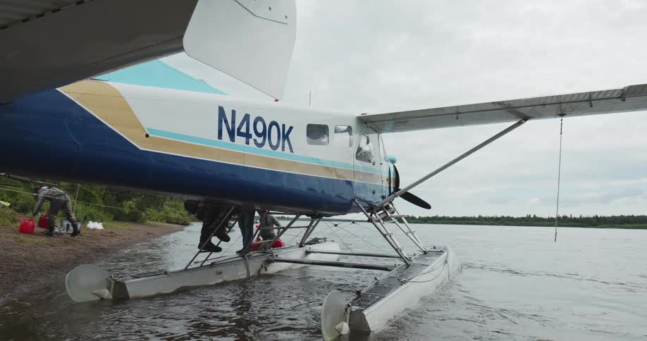 A blue and white singleengine floatplane is moored to a shoreline on a body of water as three mans step onto the floats and load equipment in a natural wilderness setting