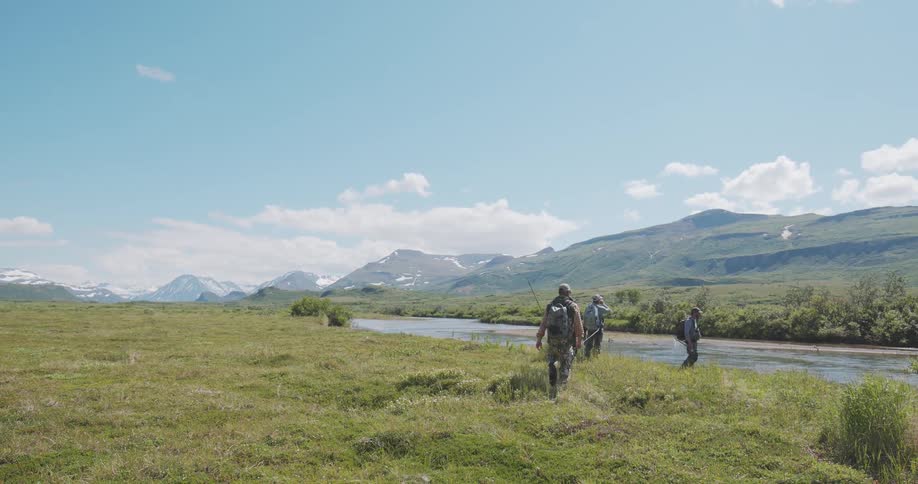 People cross grassy field near river in mountainous region under blue sky